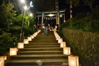 石段の両脇に明かりを灯した灯篭が並び、鳥居の奥に進む人々が見える夜の神社参道の写真
