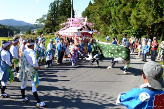 多くの観客や人々が見守る中、道路上で獅子舞と山車が向かい合いながら祭りの演舞を行っている写真