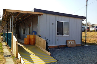 グレーの外壁に正面に窓がある平屋の建物の壁に「のぞみ福祉作業所」と書かれた木製看板が立てかけられている写真