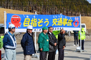 オクトパス君のイラストと「目指そう 交通事故ゼロ」と書かれた横断幕の前に人々が立っている、交通安全宣言式の写真