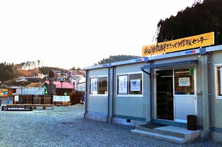 仮設のプレハブ建物に「南三陸町復興まちづくり情報センター」の看板が掲げられ、周囲には復興途中の町並みや山の斜面が広がっている南三陸町復興まちづくり情報センターの外観写真