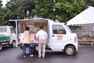 停まっている白い軽トラックの荷台を改造して店舗にした移動販売車の荷台の横が開き、その前で男性と女性が準備をしている後ろ姿を写した写真