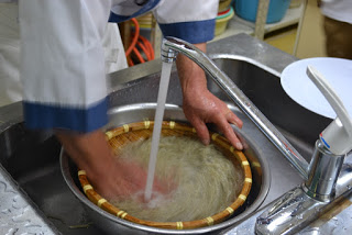 キッチンの流し台で、ボウルと茹で上がった麺が入った丸い竹ざるを蛇口からの流水で洗っている人の手元が写っている写真