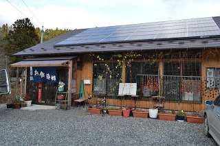 ソーラーパネルが屋根に設置された木造の建物の前に鉢植えの植物が並び、左側にある入り口には「お食事処」と書かれた暖簾が掛かっている、峠の茶や・松野やの外観写真