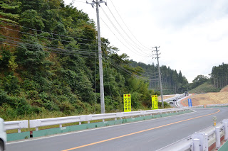 緑豊かな丘陵地帯を通る道路が写されており、両側にはガードレールと電柱が並び、奥にはカーブを描いて開けた造成地のような場所へと続いている、国道45号の写真