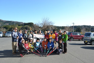 背景には木々や山が見える駐車場で、「NW体験会」と書かれた旗を持つ人を中央に、複数の参加者が屋外用の服装でウォーキングポールを持って並んでいる集合写真