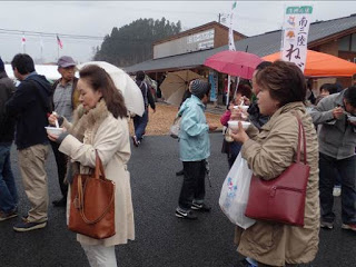 屋外で大勢の人達が傘をさしながら食べ物を食べている様子を写した南三陸グルメ笑顔まつりの様子の写真