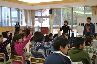 劇団四季の方々数名が教室の前に立ち、多くの子どもたちが椅子に座っていて、数人の子どもが元気よく手を挙げている様子の写真