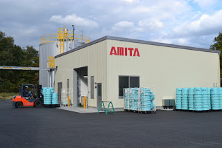 白い建物の外に「AMITA」という社名が赤く書かれており、手前には青い容器が多数並べられ、フォークリフトが作業している写真