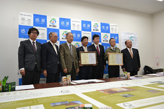 左から2番目と4番目の男性が協定書を持ち7名の関係者の人たちが並んで記念撮影をしている写真