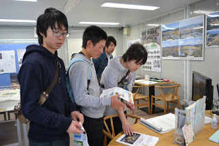 屋内の説明スペースで、数人の若者が資料を手に取りながら防災や復興に関するパネルを見学している写真