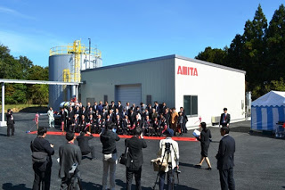 スーツ姿の多くの人々が白い建物の前で集合写真を撮影しており、建物には「AMITA」と赤い文字で書かれていて、後方には工場の設備らしきものも見える写真