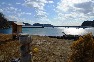 海を臨む高台の広場に、木の像と木製の小さな祠が立っている写真