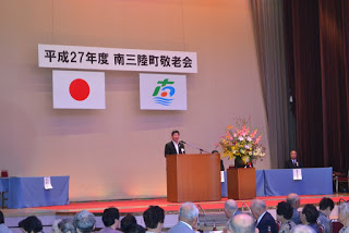 「平成27年度 南三陸町敬老会」の式典で、国旗と南三陸町町旗の下、演台に立った町長が挨拶をしている写真