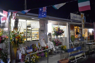 夜の屋外で、花に囲まれたお店の外観が明るく照らされており、店頭には開店祝いの花が飾られ、上空には国旗のガーランドが飾られているオーイング菓子工房Ryoの開店の風景の写真