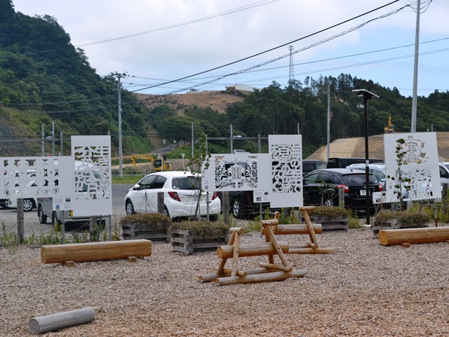 複数の車が停まった駐車場の手前にある広場に木のベンチが置かれ、5枚の白い切り絵パネルが展示されている野外展示スペースの写真