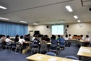 教室の中で多くの人が長机に座り、前方に設置されたスクリーンを見ながら講師の説明を聞いて講義を受けている様子を写した写真