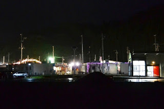 夜の暗闇の中、白っぽい外壁の建物が並ぶさんさん商店街から、やぐらに吊るされた提灯や建物の照明がやわらかく周囲を照らしている様子を遠くから写した写真