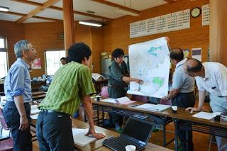 会議室で5人の男性が長机の周りに立ち、男性2人が広げた大きな地図を見ながら話し合っている様子の写真