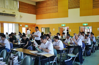 広い会場に数多く並んだ長机の席に多くの参加者が座っている写真