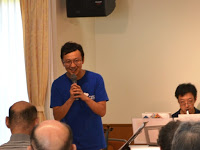 明るい部屋でマイクを握り、聴衆に向かって笑顔で話しかけている青いTシャツを着た男性を撮影した写真