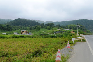 舗装された道路の左側に田畑が広がり、奥には木々に覆われた山々が見える緑豊かな景色の中、田畑の中に小さな木造の家が建っている写真