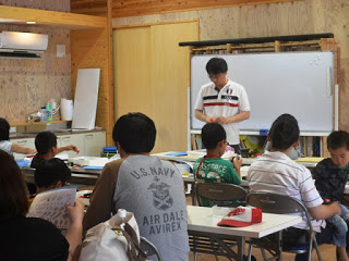 猪狩憲一さんが講師として話している教室内の風景で、参加者の皆さんが机に向かって座り、話を聞いている様子が写っている写真