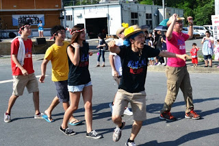 頭に紅白のねじり鉢巻きを巻いたカラフルなTシャツを着たチームが、笑顔で楽しそうに踊りながら道路を練り歩いている写真