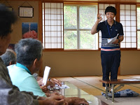 和室の畳の上に設置された長机の席に座って話を聞いている参加した高齢者に向かって、紺色のシャツを着たスタッフが紙を持ちながら説明している写真