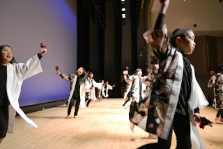 舞台の上で両手に鳴子を持ち、白と黒の衣装を着た子供たちが両手を広げて踊っている様子の写真