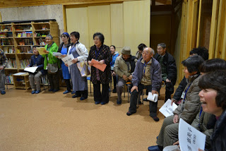 室内で高齢の男女が椅子に座って集まっており、前に立った4人の女性が資料を手に挨拶の練習をしている様子を写した写真