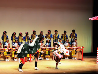 舞台の上で法被を着た子どもたちが横一列に並び、笛や太鼓の演奏に合わせて、手前で獅子舞と踊り手が演舞している写真