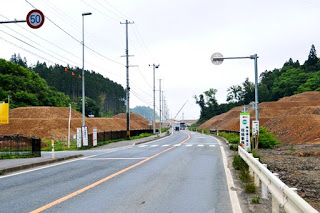左側に向かって緩やかにカーブしている道路を車やトラックが走行し、両側に看板が設置され盛り土が広がっている工事中の写真