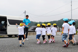 駐車場で黄色と青の帽子をかぶった複数の子供たちが、白いバンの前に立つ長谷部選手に駆け寄っている写真