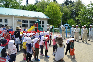 保育所の庭に赤白帽をかぶった園児たちと保育士たちが整列しており、作業服を着てヘルメットをかぶった男性たちが園児と向かい合って立っている写真