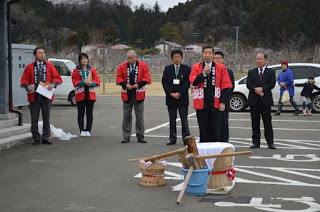 駐車場に準備された杵と臼の後ろに立った赤い法被を着た男性がマイクで話をしており、背後に同じ赤い法被を着た男女3人とスーツの男性3人が並んで立っている写真