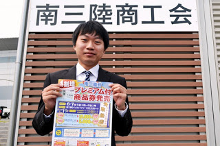 「南三陸商工会」と書かれた看板の前で、スーツ姿の男性がプレミアム付き商品券のチラシを両手で持って見せている写真