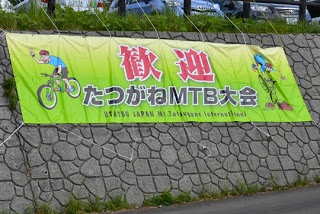 赤い文字で「歓迎」、黒と白の文字で「たつがねMTB大会」と書かれており、マウンテンバイクに乗るイラストが描かれている黄緑色の横断幕が、石垣の上に取り付けられている写真