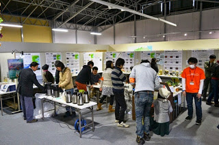 室内のイベント会場で人々がテーブルを囲み、壁に貼られた展示パネルを見たり話したりしている様子の写真