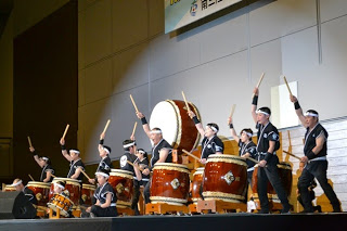 舞台の上で右手を上にあげ太鼓の演奏をしている人達を写した写真