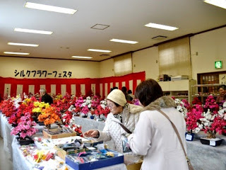 明るい室内で、色とりどりのシルクフラワー(造花)が並べられたテーブルがいくつもあり、多くの人々がそれを見ている様子の写真