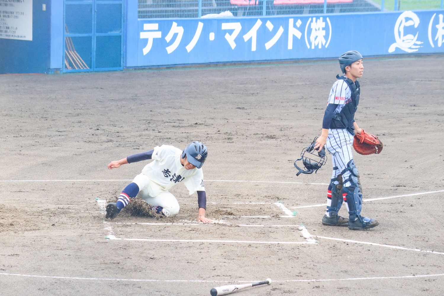 ヘルメットをかぶった白いユニフォームの選手が本塁にスライディングして得点が入る中、キャッチャーはミットと面を持ち、ボールの行方を目で追い立ち上がっているシーンの写真