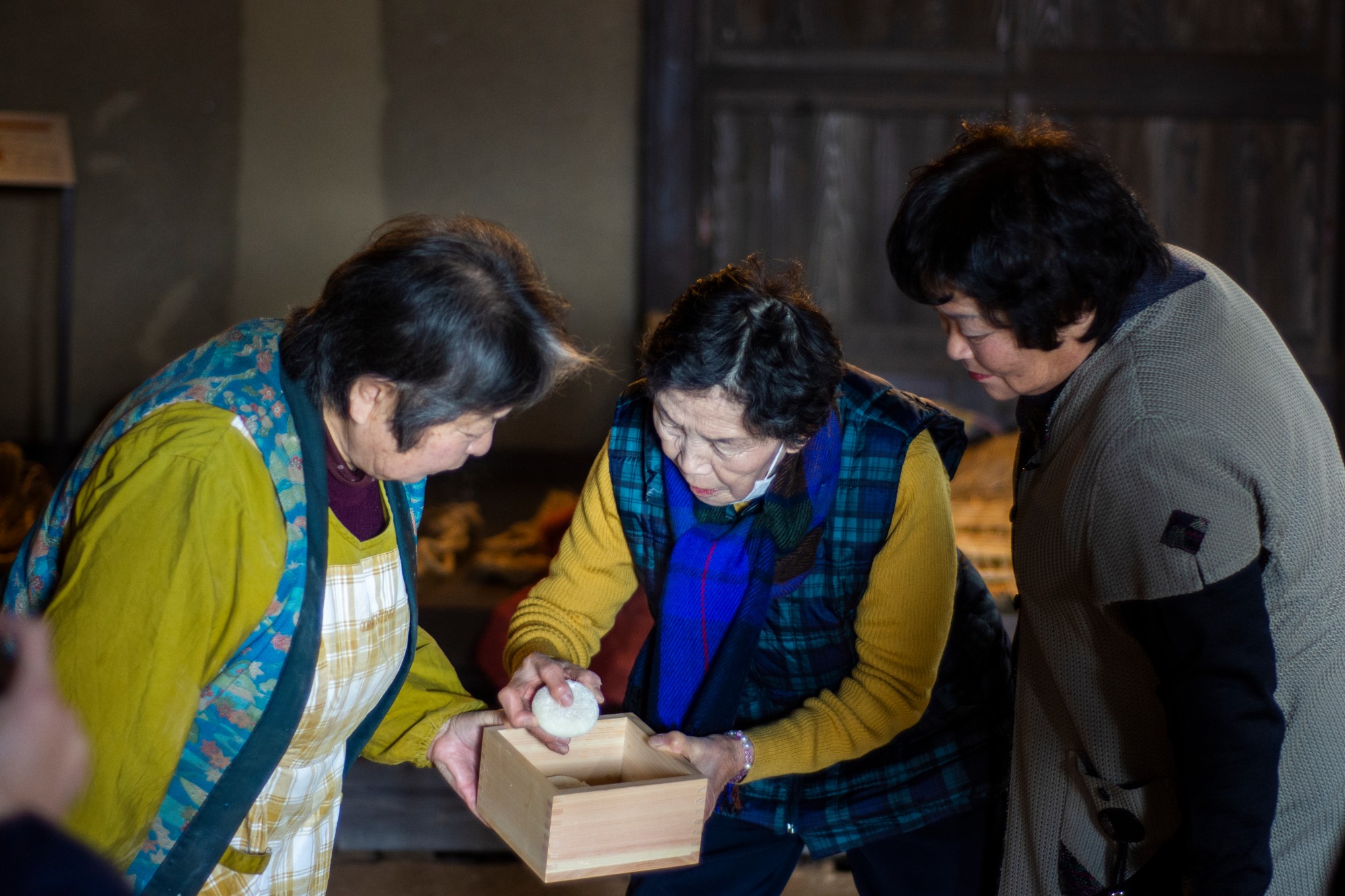 3人の女性が木製の箱を囲み、そのうちの1人が箱の中に白い餅を入れている様子が写っている写真