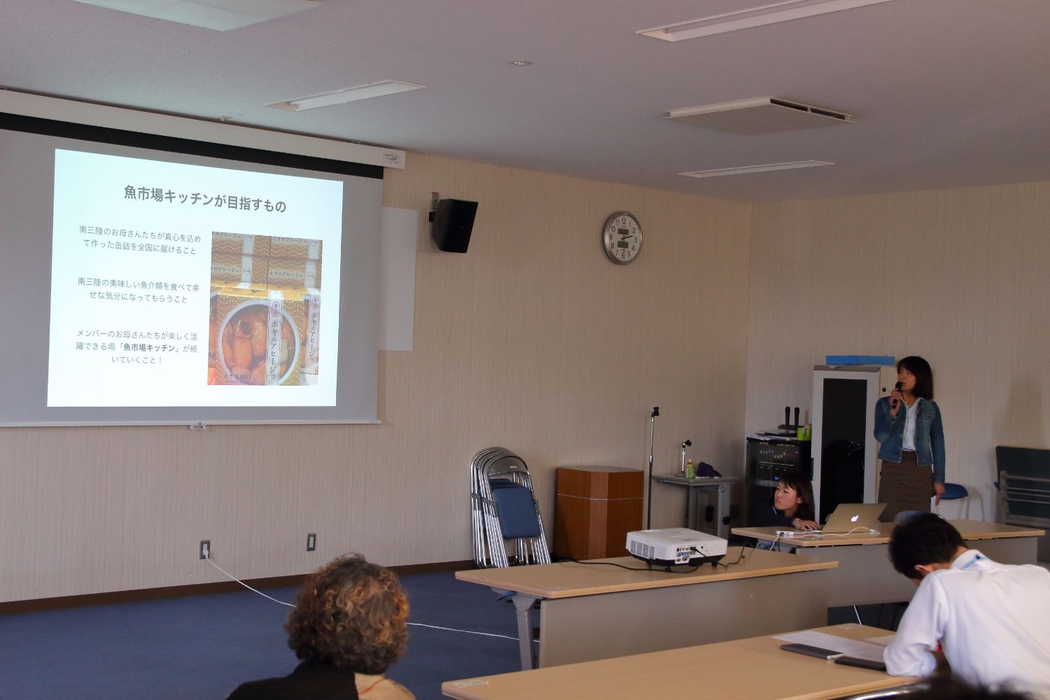 「魚市場キッチンが目指すもの」というタイトルの資料が映されたスクリーンが設置された会議室で、右側に立つ女性が話をしている様子を写している写真