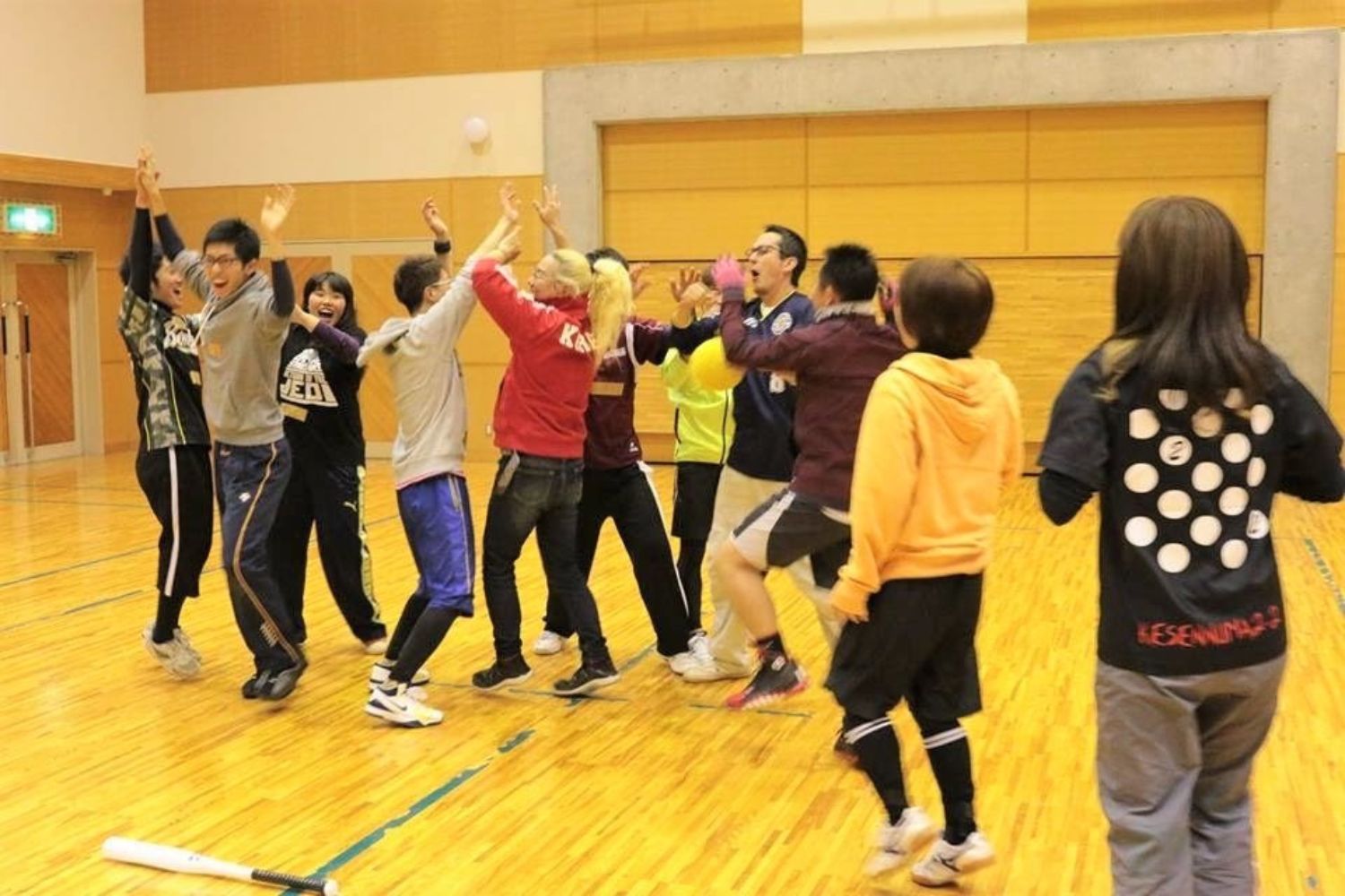 体育館の中で複数の人物が円になって立ち、手を挙げたりジャンプしたりハイタッチをしている様子が写されている写真