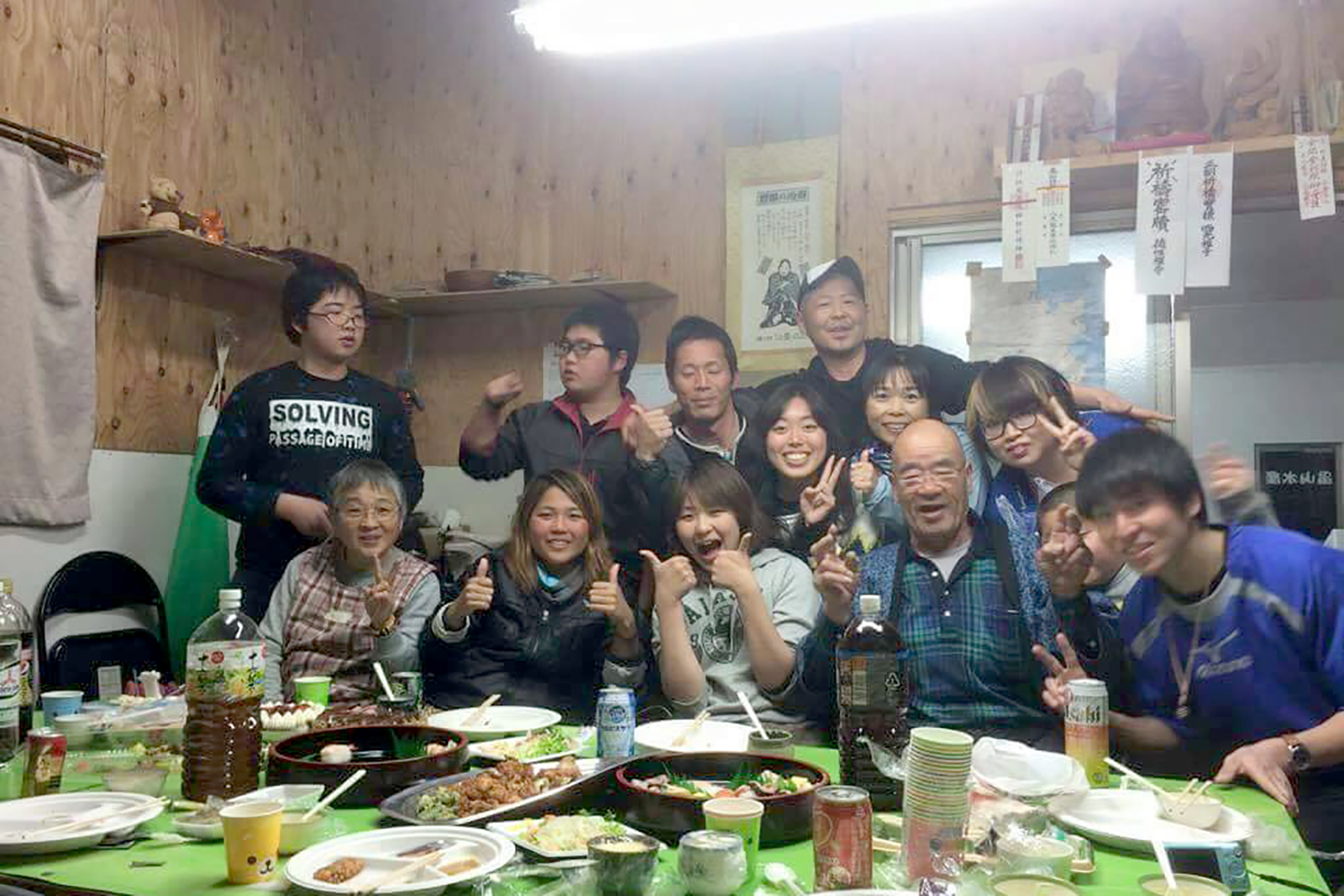 所狭しと料理や皿が並ぶテーブルの後ろで、たくさんの人が笑顔やピースサインを見せて集まっている写真