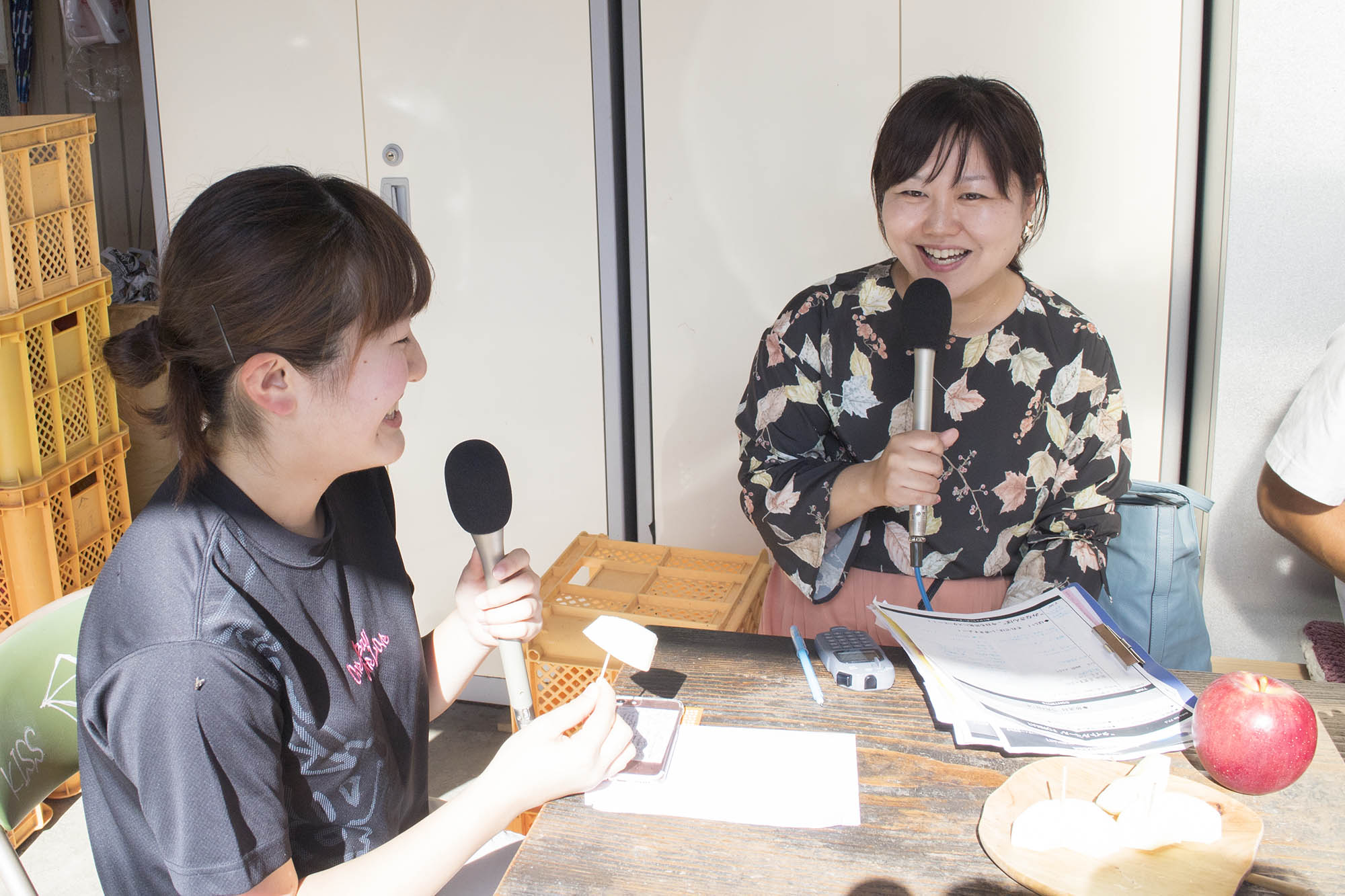 赤いりんごと、切ったりんごが盛られた皿が置かれたテーブルに座り、向かい合って笑顔でインタビューをしている女性リポーターと大沼さんの写真