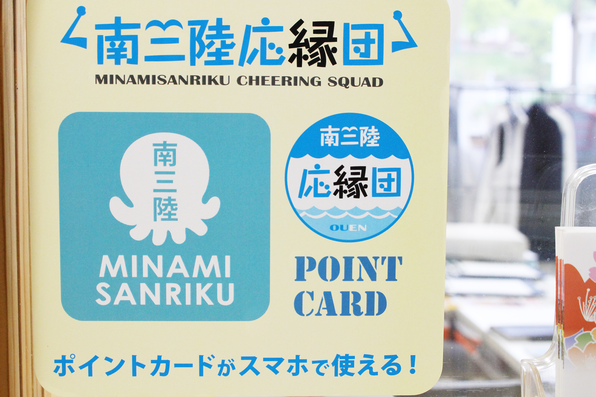店のガラス窓に貼られた「南三陸応援団 MINAMISANRIKU CHEERING SQUAD」と書かれたポスターがあり、中央には白いタコのロゴと「MINAMI SANRIKU」と書かれたマーク、その右に「POINT CARD」と書かれた円形ロゴがあり、下に「ポイントカードがスマホで使える!」と書かれているステッカーの写真