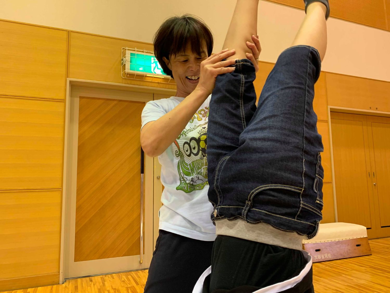 体育館で逆立ちの練習をしている子どもの足を笑顔で支えている女性の写真