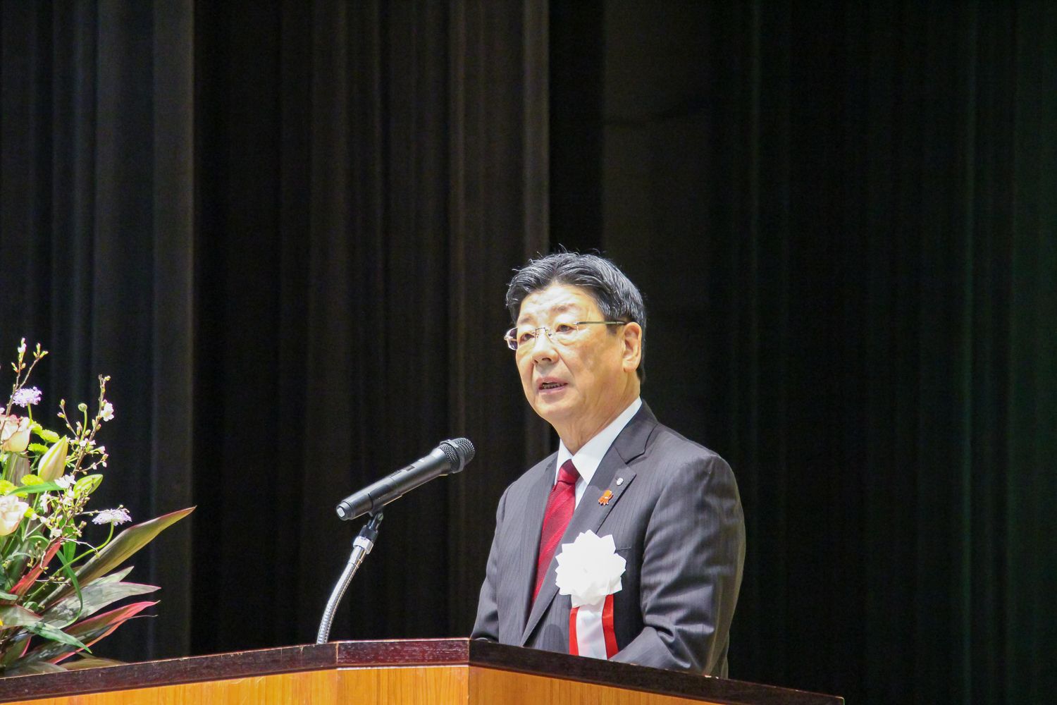 左胸に胸章リボンをつけている佐藤仁町長が壇上で話をする様子の写真